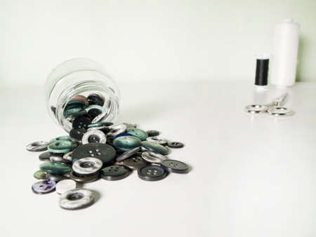 Glass jar full of clothing green, gray and black buttons, thread and scissors on white background. Sewing and craft concept.の写真素材