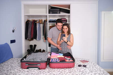 Young caucasian couple looking at the smartphone smiling, preparing a travel. Passports and sun glasses on the bed. Packing concept.の写真素材