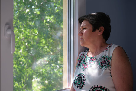 Sad, worried, thoughtful mature woman looking through the window. Sadness and quarantine concept.