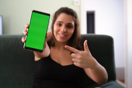 Young caucasian woman pointing at her smartphone with green screen. Chroma key.