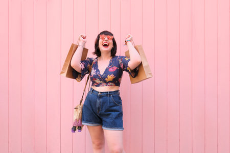 Young caucasian woman with sunglasses exited holding shopping bags over pink background. Happiness...