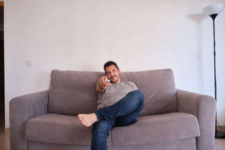 Portrait of a relaxed young man sitting on a sofa watching television and changing channels with the remote control on his hands.の写真素材