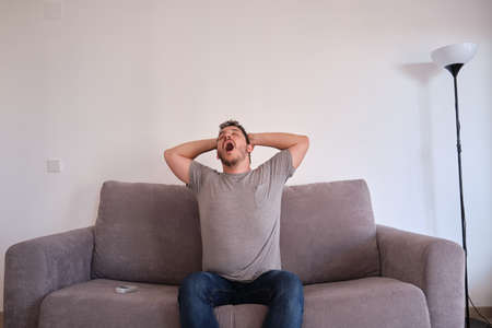 Portrait of a bored and tired young man sitting on a sofa yawning and stretching. Fatigue and tiredness concepts.の写真素材