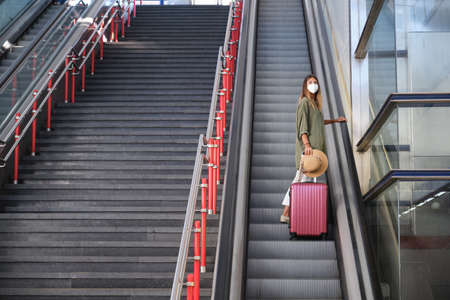 Young woman wearing face mask, and carrying a suitcase and a hat on the escalators. Travel concept.の写真素材