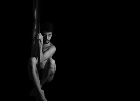 Dramatic dark portrait of a young man hanging from a pole dance bar with his eyes closed. Dark background studio shot. Black and white image.の写真素材