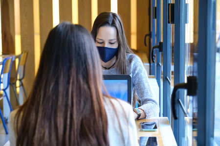 Two female students wearing face masks working on their computers in a restaurant. New normal in...