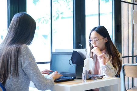 Two female students wearing face masks working on their computers in a restaurant. New normal in...