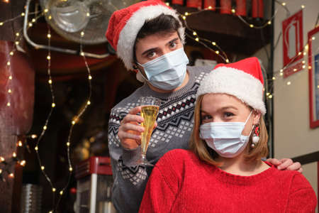Young attractive couple wearing face mask and red Santa Claus hats at a Christmas party in a bar. Coronavirus new normal.の写真素材