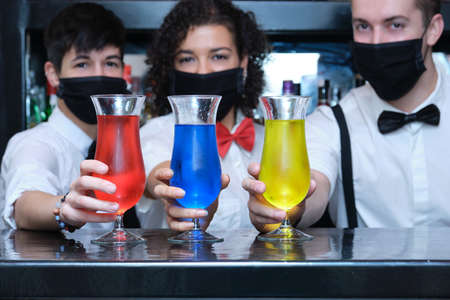 Three waiters wearing protective face mask grabbing three colorful cocktails in a pub. New normal in pubs.の写真素材