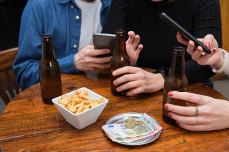 Unrecognizable three friends seeing something on their smartphone, with drink, snacks and money on the table. Bar routine concept.の写真素材