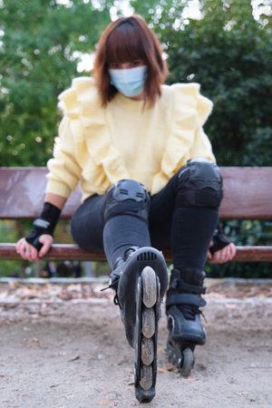 Young caucasian woman wearing face mask is sitting in a bench with her roller skates on. Outdoor...