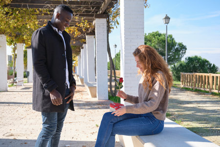 African black man giving a rose and a heart shape box with sweets to his redhead caucasian...