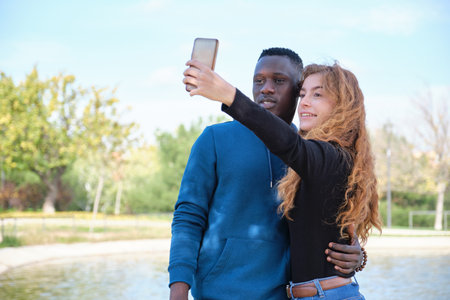 African black man and redhead caucasian woman smiling and taking a selfie with their smartphone in a park. Young multiracial couple portrait.の写真素材