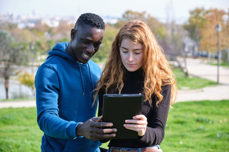 African black man and redhead caucasian woman looking at their tablet in a park. Young multiracial...
