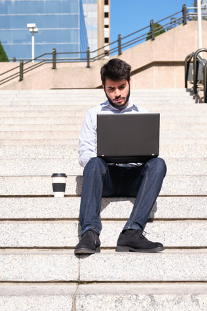 Young business man using his laptop, with a coffee cup, sitting on stairs. Businessman in a urban street of a city.の写真素材