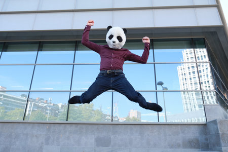 Young business man wearing a panda head mask, jumping in front of the a corporate building. Bizarre businessman.の写真素材