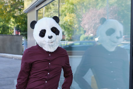 Young business man wearing a panda head mask reflected on a glass of a corporate building. Bizarre businessman.の写真素材