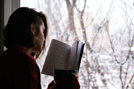 Young woman holding a hot coffee or tea cup, reading a book by the window in a snowy winter day. Cozy at home concept.の写真素材