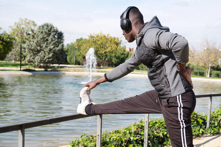 African black man with headphones stretching his leg in a park. Fitness and sport outdoor.