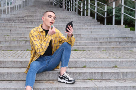 Portrait of a handsome young man, applying lipstick in front of the mirror, wearing black eyeshadow, holding a makeup brush. Non binary androgynous guy.の写真素材