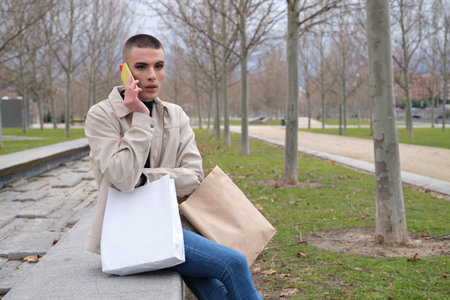 Handsome young man wearing make up, worried speaking on his smartphone, carrying shopping bags. Non...