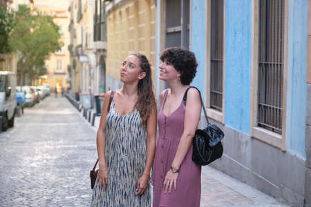 Beautiful lesbian couple wearing long dresses looking at something in the street. Young woman friends.の写真素材
