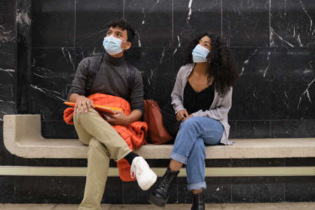 Young latin couple wearing protective face mask sitting at the metro station waiting for the train. New normal at public transport.の写真素材