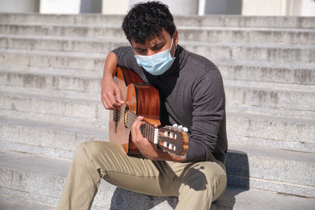 Latin young man wearing protective face mask playing guitar sitting on stairs. Playing guitar...