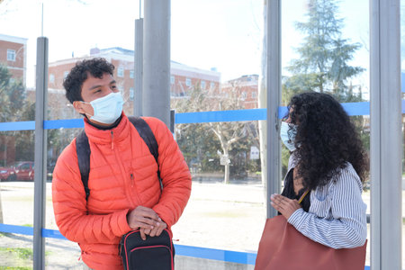 Young latin couple wearing protective face mask and carrying guitar case looking at the bus,...