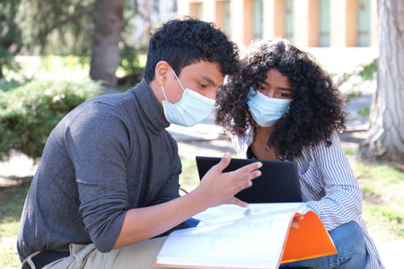 Two latin students wearing protective face mask studying together sitting on a bench outdoor. New...