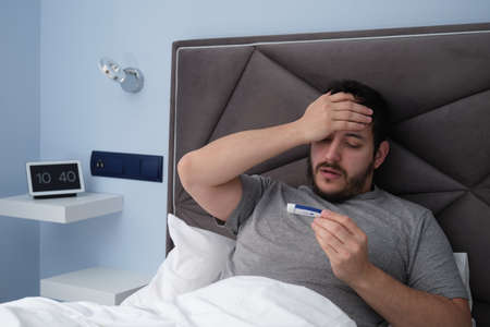 Young latin man sick in bed with a thermometer in his hand. Digital clock at the background.の写真素材