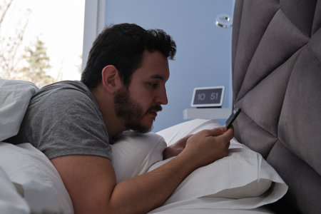 Young latin man with his smartphone in bed late in the morning. Digital clock at the background.