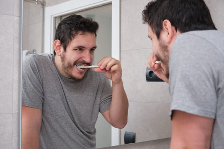 Young latin man brushing his teeth in front of the mirror in the bathroom at morning. Eco-friendly...