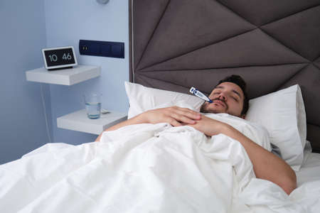 Young latin man sick in bed with a thermometer in his mouth. Digital clock, water and pills at the background.の写真素材