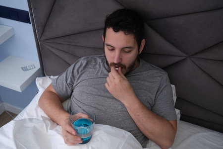 Young latin man ill in bed taking a pill and a glass of water.