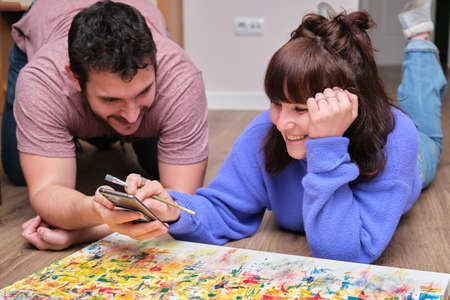 Young couple smiling and seeing something on the smartphone while she is painting on canvas with oil paints at home. Artist drawing laying on the floor.の写真素材