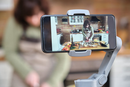 Young woman cooking stuffed aubergines and recording a video with her smartphone for her cooking...