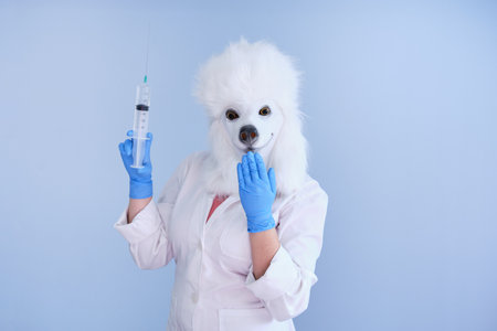 Young woman in a latex dog head mask and white coat holding a big syringe and giggle on a blue background. Doctor medical veterinary concepts.の写真素材
