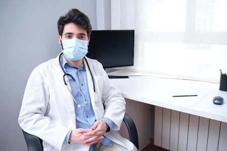 Portrait of professional confident young male doctor in medical mask and white coat, stethoscope over neck, sitting on his desk during pandemic.の写真素材