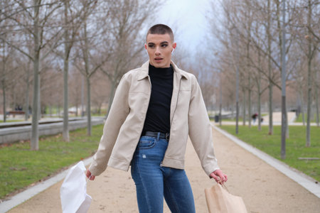 Young man wearing make up spinning around with shopping bags and listening music in a park. Queer...