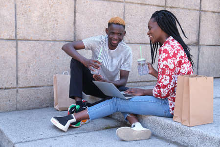 Young african couple laughing, checking a laptop, drinking refreshments and sitting on stairs after going shopping. City life concept.の写真素材