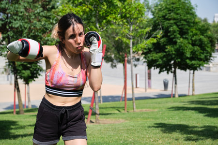Young woman in boxing gloves hitting and looking at camera. Muay thai, sport outdoors.の写真素材
