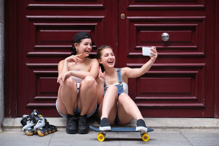 Two women laughing and taking a selfie, holding a skateboard and inline skates in a city. Friends,...