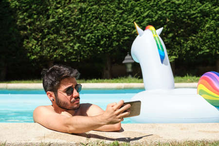 Young man wearing sunglasses, taking a selfie in a swimming pool. Unicorn inflatable ring at background. Summer concept.の写真素材