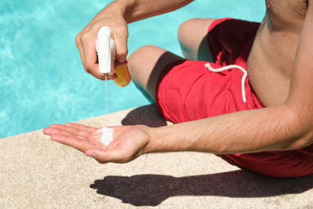 Unrecognizable young man applying sunscreen sitting at the edge of the swimming pool. Summer concept.の写真素材