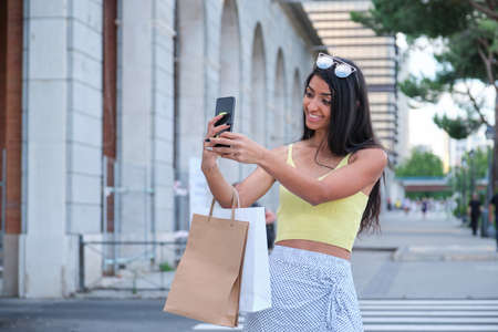 Young latin woman taking a selfie and holding shopping bags at street.の写真素材