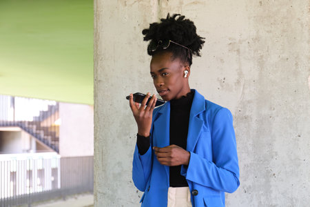 Portrait of smiling young african woman wearing wireless earbuds, recording an audio note with her smartphone, wearing blue jacket suit and sunglasses over a concrete wall.の写真素材