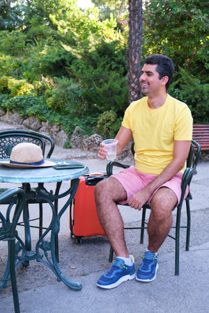 Tourist resting drinking a refreshment and sitting on a bar summer terrace with his suitcase and a hat.の写真素材