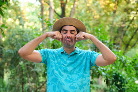 Man pretending that is crying because summer vacation has finished, with a straw hat and a blue pole shirt in nature.の写真素材