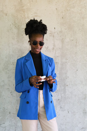 Portrait of smiling young african woman putting on her wireless earbuds, with a blue jacket suit and sunglasses over a concrete wall.の写真素材